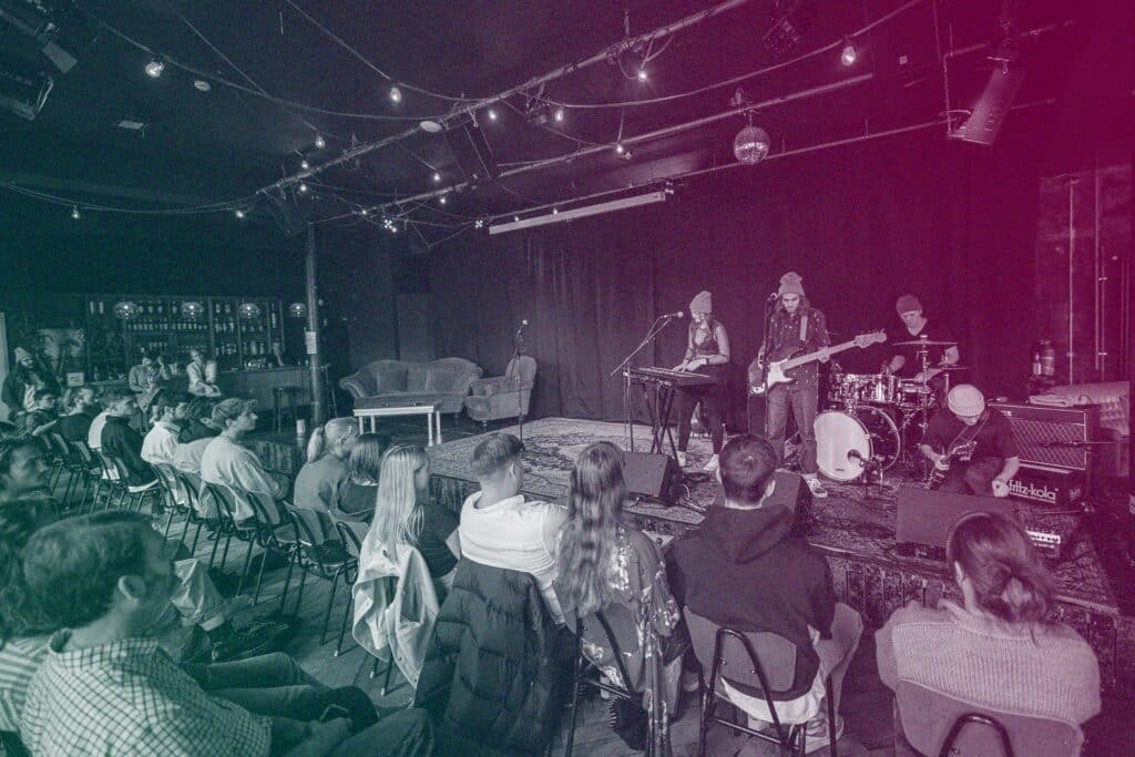 A band plays on stage to an audience. The band comprises of a keyboard player, a vocalist and guitarist, and a drummer.